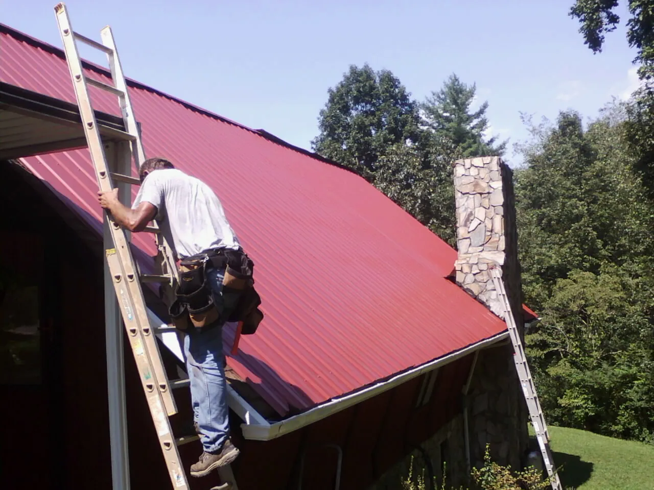 Roofer on a ladder beside a red metal roof and stone chimney — Soffit & Fascia Repair by Roofing Brothers in Newnan